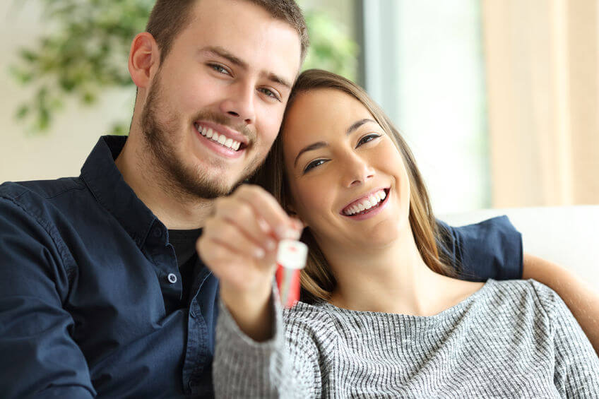 happy couple of owners showing house keys to the camera sitting on a sofa in the living room at home